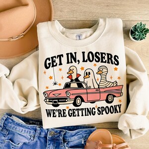 K&ouml;nnte beinhalten: Cremefarbenes Sweatshirt mit dem Text "GET IN, LOSERS WE'RE GETTING SPOOKY" und einer Grafik von drei Cartoon-Enten in einem rosa Auto. Die Enten sind als Vampir, Geist und Mumie verkleidet. Sterne und eine Spinne.