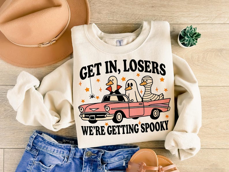 K&ouml;nnte beinhalten: Cremefarbenes Sweatshirt mit dem Text "GET IN, LOSERS WE'RE GETTING SPOOKY" und einer Grafik von drei Cartoon-Enten in einem rosa Auto. Die Enten sind als Vampir, Geist und Mumie verkleidet. Sterne und eine Spinne.