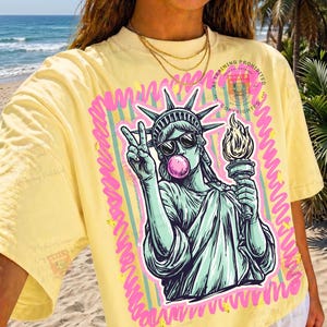 Puede incluir: Camiseta amarilla con un gr&aacute;fico de la Estatua de la Libertad con gafas de sol y soplando una burbuja de chicle, haciendo el signo de la paz. El gr&aacute;fico est&aacute; enmarcado con l&iacute;neas rosas y verdes. El texto "AT TRAINING PROHIBITED" es visible.