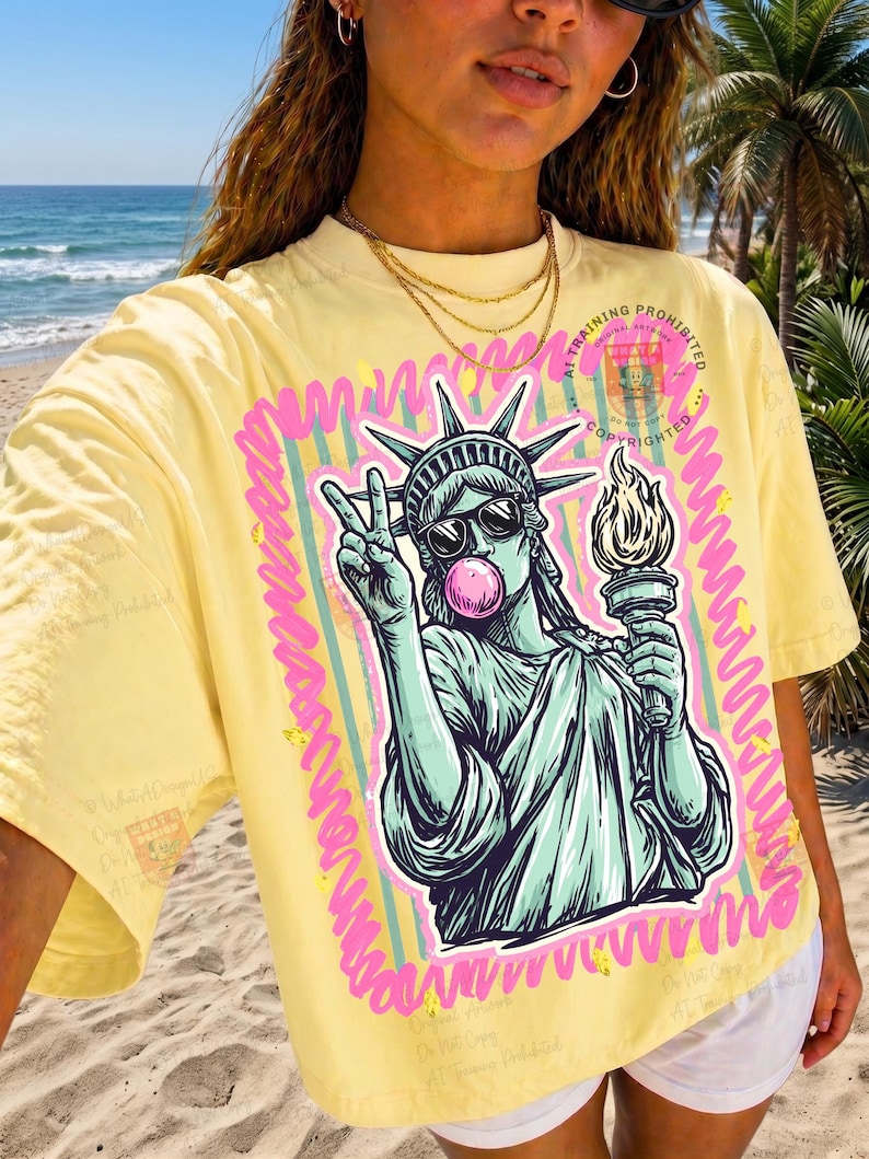 Puede incluir: Camiseta amarilla con un gr&aacute;fico de la Estatua de la Libertad con gafas de sol y soplando una burbuja de chicle, haciendo el signo de la paz. El gr&aacute;fico est&aacute; enmarcado con l&iacute;neas rosas y verdes. El texto "AT TRAINING PROHIBITED" es visible.