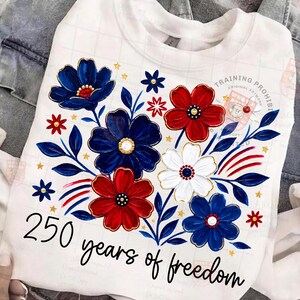 May include: White sweatshirt with a floral design in red, white, and blue. The flowers are accented with gold trim and surrounded by blue leaves and small gold stars. The text "250 years of freedom" is written in black script.
