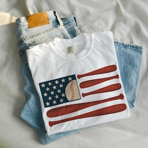 May include: White t-shirt with a baseball-themed graphic. The design features a baseball, stars, and baseball bats arranged to resemble a flag. The shirt is laid on top of a pair of blue jeans.