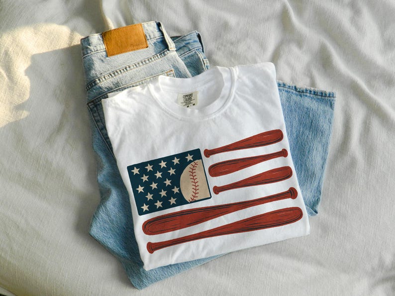 May include: White t-shirt with a baseball-themed graphic. The design features a baseball, stars, and baseball bats arranged to resemble a flag. The shirt is laid on top of a pair of blue jeans.