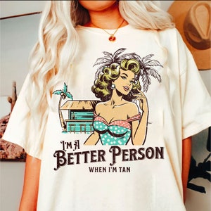 May include: White t-shirt with a retro cartoon illustration of a woman in a swimsuit and a palm tree. The text "I'm a Better Person When I'm Tan" is printed on the shirt.