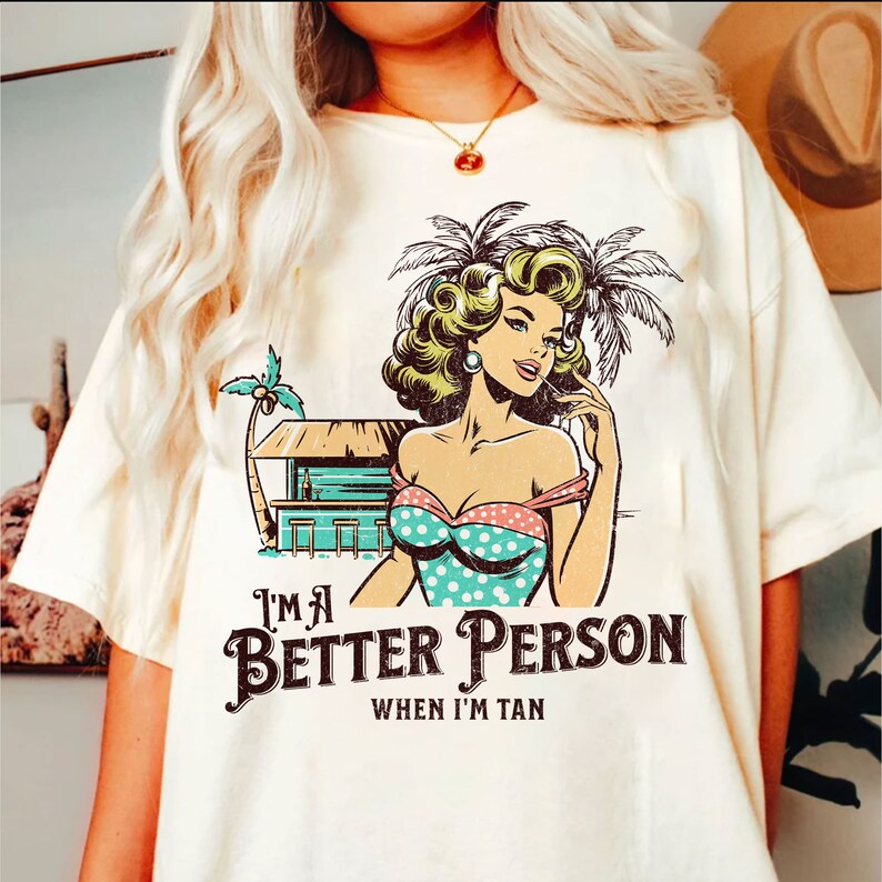 May include: White t-shirt with a retro cartoon illustration of a woman in a swimsuit and a palm tree. The text "I'm a Better Person When I'm Tan" is printed on the shirt.