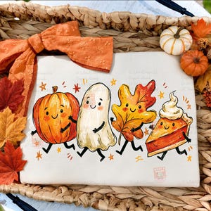 May include: A white t-shirt featuring a colorful graphic of a smiling pumpkin, a ghost, an autumn leaf, and a slice of pumpkin pie, all with arms and legs. The shirt is decorated with an orange eyelet fabric bow and surrounded by fall foliage.