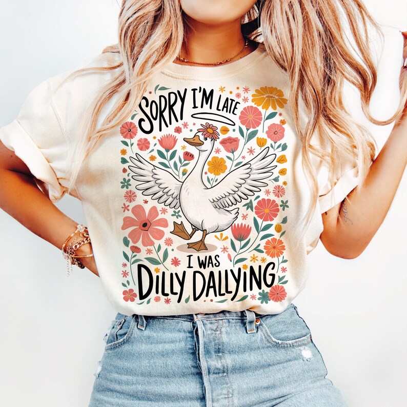 May include: A white t-shirt featuring a floral design and a cartoon goose. The text on the shirt reads "Sorry I'm Late I Was Dilly Dallying".