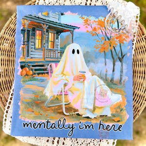 K&ouml;nnte beinhalten: Ein hellblaues T-Shirt mit einer grafischen Illustration eines Geistes, der auf einem Stuhl vor einem Haus sitzt und einen Kaffeebecher h&auml;lt. Die Szene zeigt herbstliche B&auml;ume und K&uuml;rbisse. Der Text "mentally i'm here" ist unten aufgedruckt.