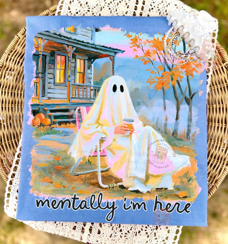 K&ouml;nnte beinhalten: Ein hellblaues T-Shirt mit einer grafischen Illustration eines Geistes, der auf einem Stuhl vor einem Haus sitzt und einen Kaffeebecher h&auml;lt. Die Szene zeigt herbstliche B&auml;ume und K&uuml;rbisse. Der Text "mentally i'm here" ist unten aufgedruckt.