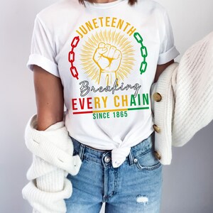 Peut inclure: T-shirt blanc avec un motif graphique repr&eacute;sentant un poing lev&eacute;, des cha&icirc;nes et le texte "Juneteenth Breaking Every Chain Since 1865".