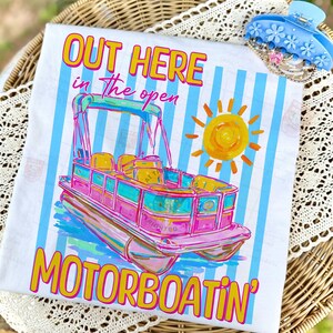 May include: White t-shirt with a colorful illustration of a pink and teal pontoon boat, a yellow sun, and the text "OUT HERE in the open MOTORBOATIN'". The shirt has blue vertical stripes and is next to a blue floral hair clip.