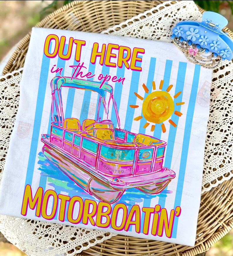 May include: White t-shirt with a colorful illustration of a pink and teal pontoon boat, a yellow sun, and the text "OUT HERE in the open MOTORBOATIN'". The shirt has blue vertical stripes and is next to a blue floral hair clip.