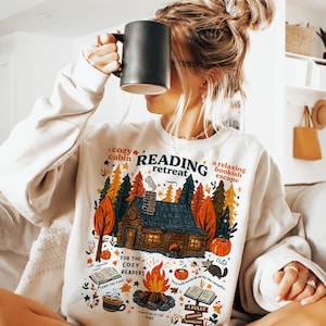 Pode incluir: Camisola creme com um gr&aacute;fico colorido de uma cabana na floresta, ab&oacute;boras e uma fogueira. O texto na camisola diz "Reading Retreat" e "For the Cozy Readers". Uma caneca preta &eacute; segurada em frente ao rosto.