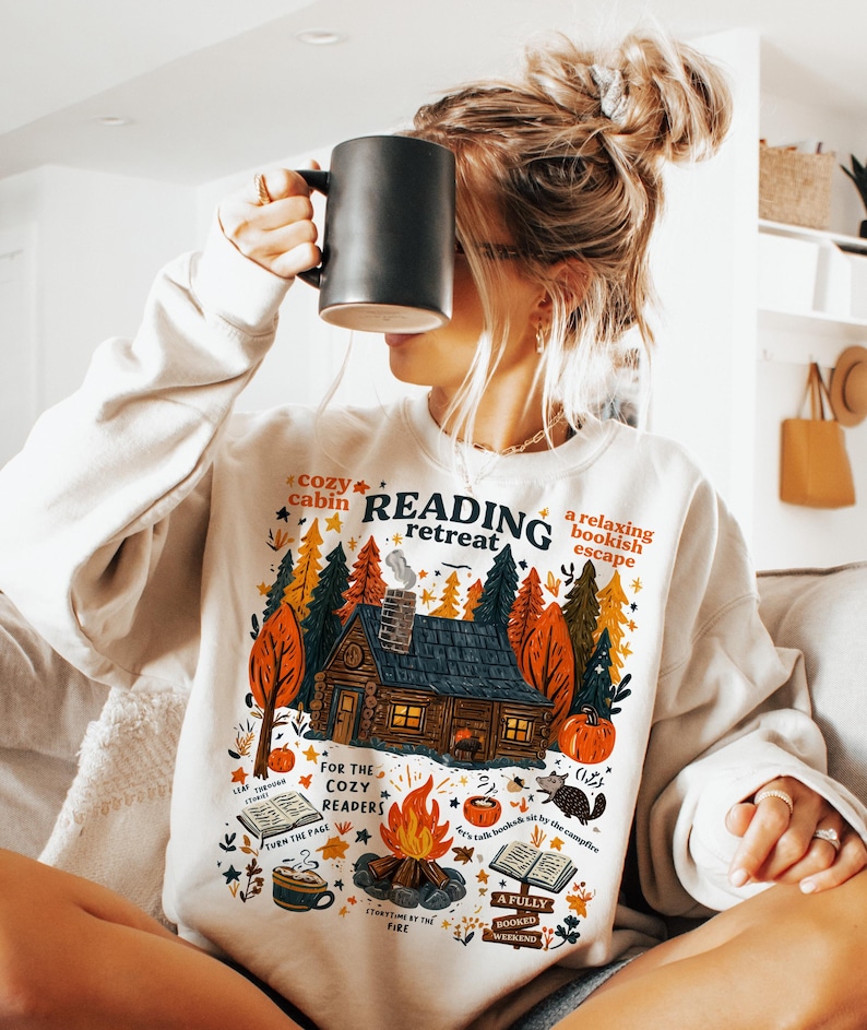 Pode incluir: Camisola creme com um gr&aacute;fico colorido de uma cabana na floresta, ab&oacute;boras e uma fogueira. O texto na camisola diz "Reading Retreat" e "For the Cozy Readers". Uma caneca preta &eacute; segurada em frente ao rosto.