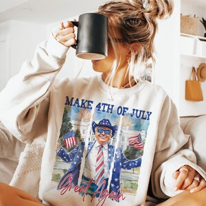 May include: A white sweatshirt with a graphic print of a person wearing a cowboy hat and sunglasses holding two American flags. The text "Make 4th of July Great Again" is printed on the sweatshirt.
