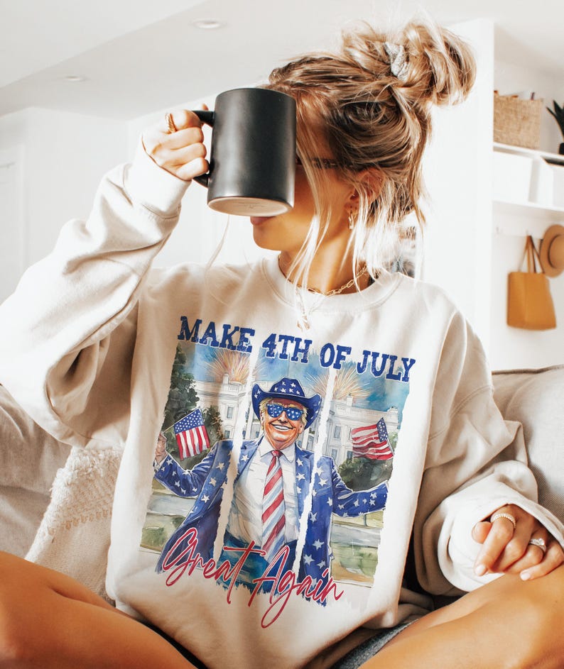 May include: A white sweatshirt with a graphic print of a person wearing a cowboy hat and sunglasses holding two American flags. The text "Make 4th of July Great Again" is printed on the sweatshirt.