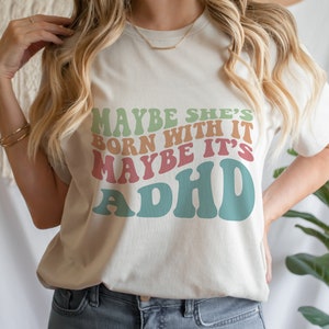 May include: Cream-colored t-shirt with the text "MAYBE SHE'S BORN WITH IT MAYBE IT'S ADHD" in retro-style lettering. The text is in shades of green, orange, pink, and blue. The shirt has a crew neck and short sleeves.