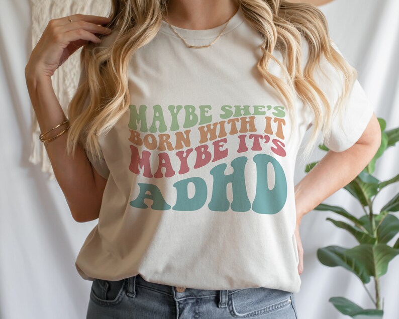 May include: Cream-colored t-shirt with the text "MAYBE SHE'S BORN WITH IT MAYBE IT'S ADHD" in retro-style lettering. The text is in shades of green, orange, pink, and blue. The shirt has a crew neck and short sleeves.