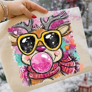 May include: A tan tote bag featuring a cartoon reindeer with yellow sunglasses blowing a pink bubblegum bubble. The reindeer wears a striped scarf and has colorful accents. The image has a winter theme.