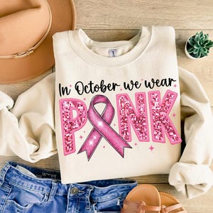 May include: A cream-colored sweatshirt with the text "In October we wear PINK" in black lettering. The word "PINK" is decorated with pink glitter and a pink ribbon.