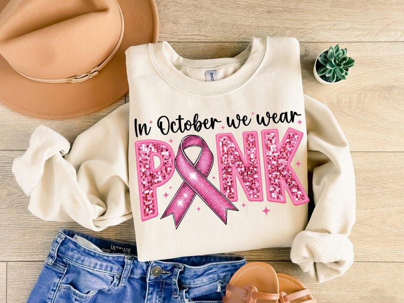 May include: A cream-colored sweatshirt with the text "In October we wear PINK" in black lettering. The word "PINK" is decorated with pink glitter and a pink ribbon.