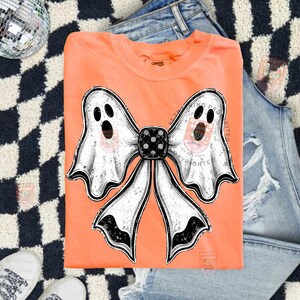 Op de afbeelding: Een feloranje T-shirt met ronde hals en een grote afbeelding van een witte strik gevormd door twee geesten met zwarte ogen en monden. Het midden van de strik is een zwarte dobbelsteen met witte stippen. Het ontwerp heeft een verweerde look.