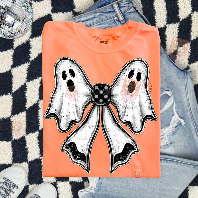 Op de afbeelding: Een feloranje T-shirt met ronde hals en een grote afbeelding van een witte strik gevormd door twee geesten met zwarte ogen en monden. Het midden van de strik is een zwarte dobbelsteen met witte stippen. Het ontwerp heeft een verweerde look.