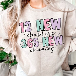 K&ouml;nnte beinhalten: Ein beiger Pullover mit dem Text "12 NEUE Kapitel 365 NEUE Chancen" in bunten Blockbuchstaben.