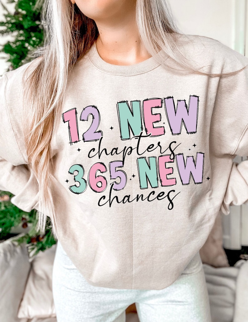 K&ouml;nnte beinhalten: Ein beiger Pullover mit dem Text "12 NEUE Kapitel 365 NEUE Chancen" in bunten Blockbuchstaben.