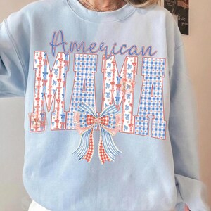 Op de afbeelding: Lichtblauw sweatshirt met de tekst "American MAMA". De letters zijn versierd met rood, wit en blauw geruit patroon en strikken. Een grote strik is onder de tekst geplaatst.