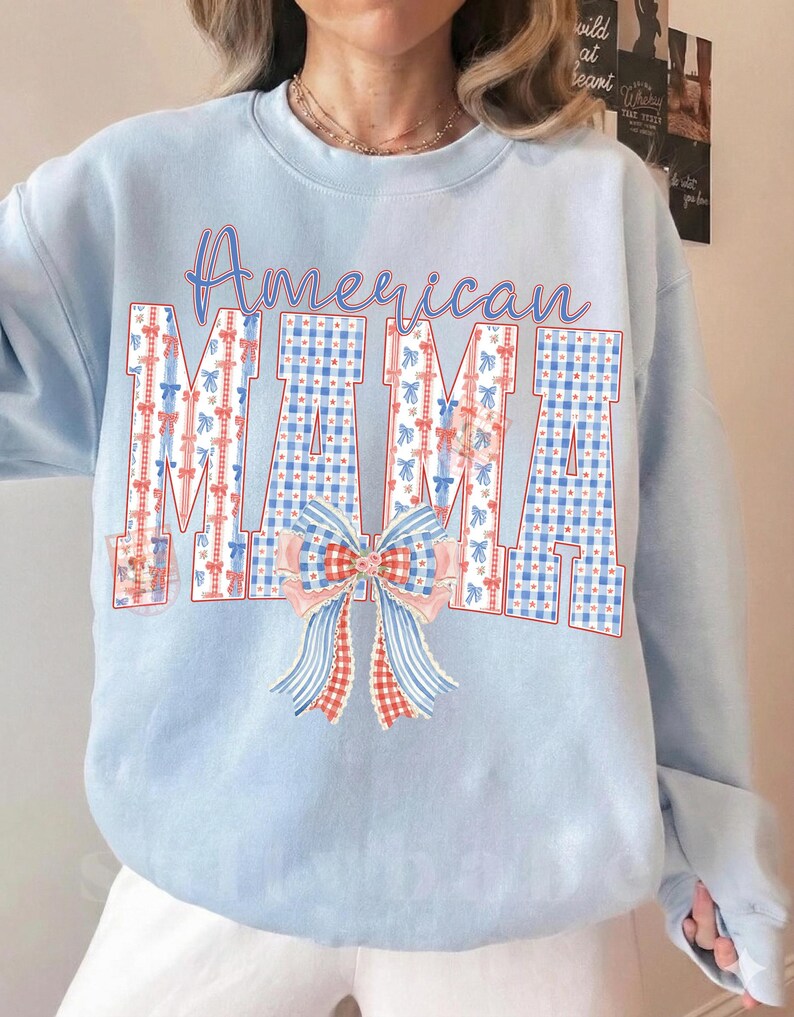 Op de afbeelding: Lichtblauw sweatshirt met de tekst "American MAMA". De letters zijn versierd met rood, wit en blauw geruit patroon en strikken. Een grote strik is onder de tekst geplaatst.