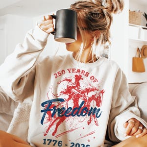 May include: Cream-colored sweatshirt with a graphic featuring a cowboy on a horse in red and blue, with the words "250 Years of Freedom" and the dates "1776-2026". The sweatshirt is a casual style.