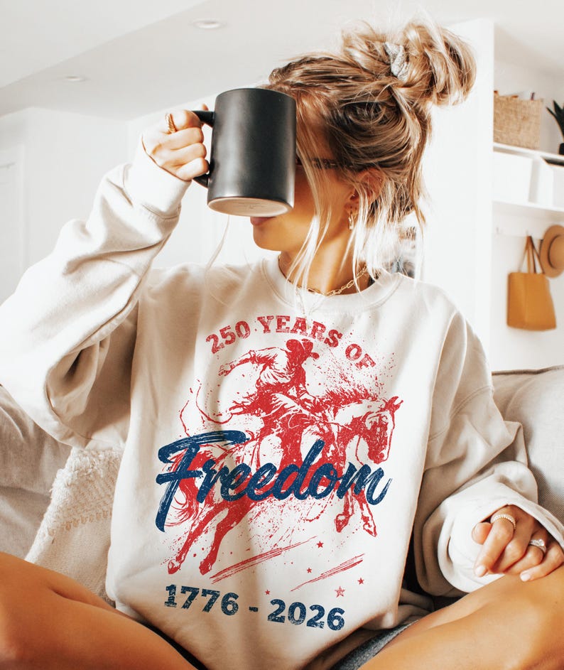 May include: Cream-colored sweatshirt with a graphic featuring a cowboy on a horse in red and blue, with the words "250 Years of Freedom" and the dates "1776-2026". The sweatshirt is a casual style.