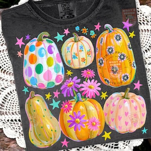May include: A charcoal gray t-shirt featuring a graphic print of five pumpkins in various shapes and colors, decorated with polka dots, floral patterns, and hearts, surrounded by colorful stars and pink and purple daisy-like flowers.