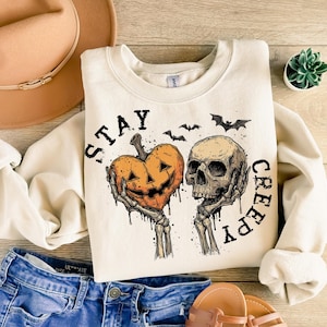 Puede incluir: Sudadera color crema con un gr&aacute;fico tem&aacute;tico de Halloween. El dise&ntilde;o presenta un coraz&oacute;n de calabaza tallada y una calavera sostenida por manos esquel&eacute;ticas, con las palabras "STAY CREEPY" en negro. Tambi&eacute;n se incluyen murci&eacute;lagos.