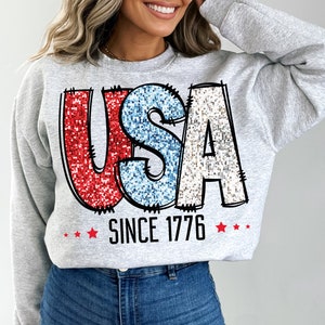May include: A gray sweatshirt with a glittery red, white, and blue "USA" graphic and the text "Since 1776" with red stars.