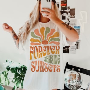 May include: White t-shirt with a retro-style graphic of a sunburst design in shades of orange, green, and yellow, with the words "FOREVER CHASING SUNSETS" in a matching font. The person is wearing a black baseball cap with white text.