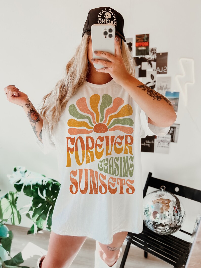 May include: White t-shirt with a retro-style graphic of a sunburst design in shades of orange, green, and yellow, with the words "FOREVER CHASING SUNSETS" in a matching font. The person is wearing a black baseball cap with white text.