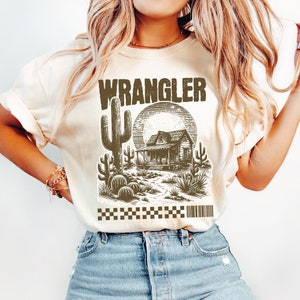 Pode incluir: Uma camiseta bege com um gr&aacute;fico marrom de uma cena do deserto com uma casa, cactos e a palavra "WRANGLER" impressa em letras marrons.