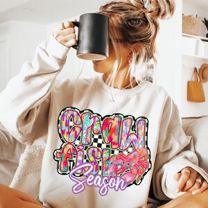 May include: A white sweatshirt with a colorful graphic design that says "Crawfish Season" with a crawfish illustration. The sweatshirt is being worn by a person with long blonde hair.