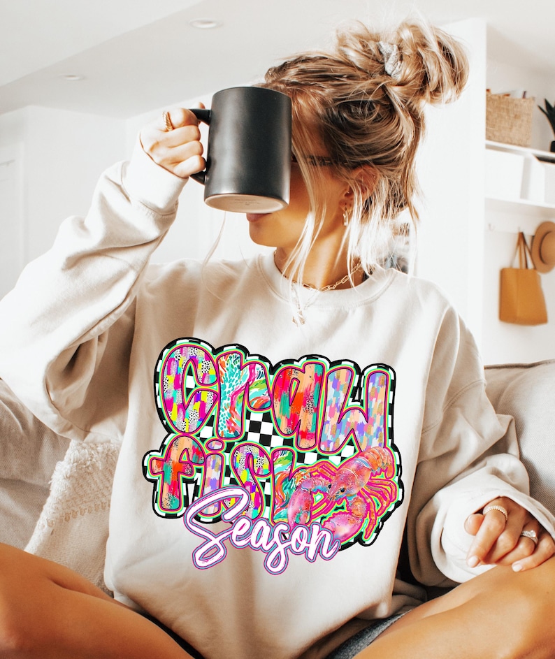 May include: A white sweatshirt with a colorful graphic design that says "Crawfish Season" with a crawfish illustration. The sweatshirt is being worn by a person with long blonde hair.