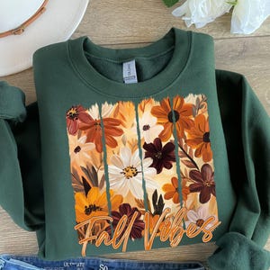 May include: A dark green sweatshirt featuring a floral design with orange, white, and brown flowers. The words "Fall Vibes" are written in an orange script across the bottom of the floral design. The sweatshirt is folded.