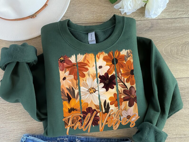May include: A dark green sweatshirt featuring a floral design with orange, white, and brown flowers. The words "Fall Vibes" are written in an orange script across the bottom of the floral design. The sweatshirt is folded.
