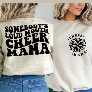 May include: A cream colored sweatshirt with black text on the back that reads "Somebody's Loud Mouth Cheer Mama". The front of the sweatshirt has a black and white pom-pom with the text "Cheer Mama" around it.