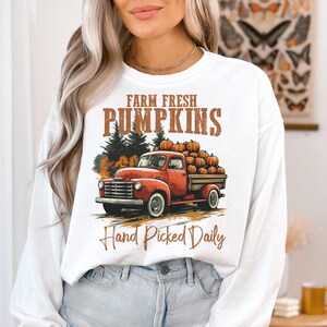 Op de afbeelding: Witte longsleeve sweatshirt met een afbeelding van een rode vrachtwagen die pompoenen vervoert. De tekst "Farm Fresh Pumpkins Hand Picked Daily" staat op de sweatshirt gedrukt.