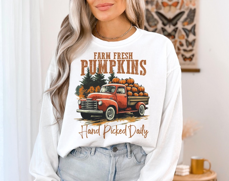 Op de afbeelding: Witte longsleeve sweatshirt met een afbeelding van een rode vrachtwagen die pompoenen vervoert. De tekst "Farm Fresh Pumpkins Hand Picked Daily" staat op de sweatshirt gedrukt.