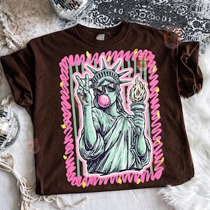 Puede incluir: Camiseta marr&oacute;n con un gr&aacute;fico de la Estatua de la Libertad con gafas de sol y soplando una burbuja de chicle. La Estatua de la Libertad sostiene una antorcha y hace el signo de la paz. El gr&aacute;fico est&aacute; enmarcado con l&iacute;neas rosas y verdes.