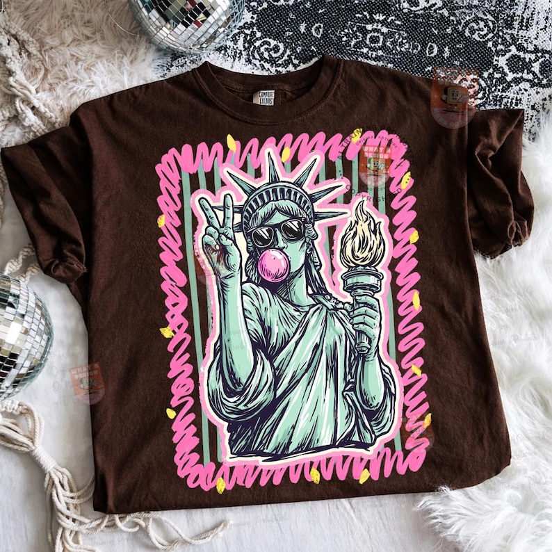 Puede incluir: Camiseta marr&oacute;n con un gr&aacute;fico de la Estatua de la Libertad con gafas de sol y soplando una burbuja de chicle. La Estatua de la Libertad sostiene una antorcha y hace el signo de la paz. El gr&aacute;fico est&aacute; enmarcado con l&iacute;neas rosas y verdes.