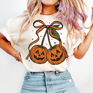 May include: Cream-colored t-shirt featuring a Halloween-themed graphic. The design showcases two orange pumpkin-shaped cherries with smiling jack-o'-lantern faces, connected by stems and a bow. Small black stars are scattered around the pumpkins.
