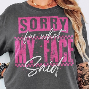 May include: Dark gray t-shirt with the text "SORRY for what MY FACE Said" in pink and white. The shirt has a vintage, worn look, with a pink checkered pattern and star accents.